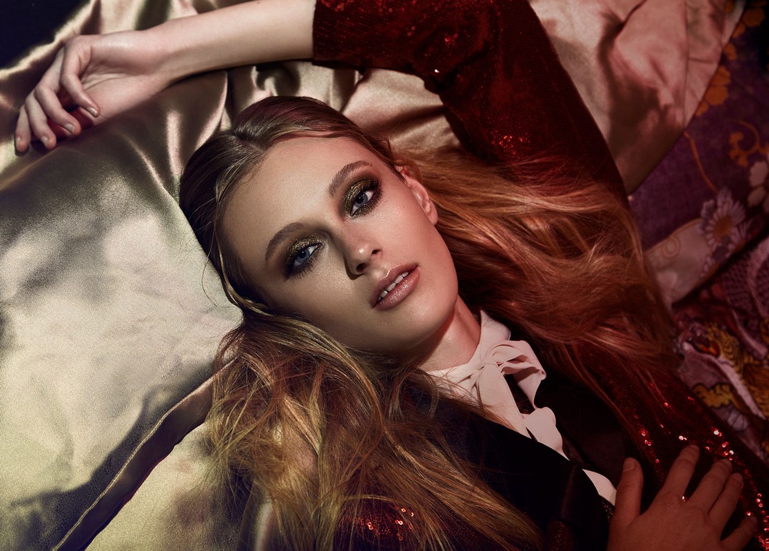 Fashion portrait of a blonde woman lying on a gold fabric, wearing a red sequined jacket with glamorous makeup and dramatic lighting.