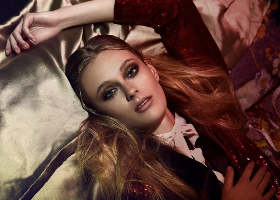 Fashion portrait of a blonde woman lying on a gold fabric, wearing a red sequined jacket with glamorous makeup and dramatic lighting.