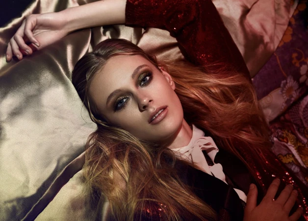 Fashion portrait of a blonde woman lying on a gold fabric, wearing a red sequined jacket with glamorous makeup and dramatic lighting.