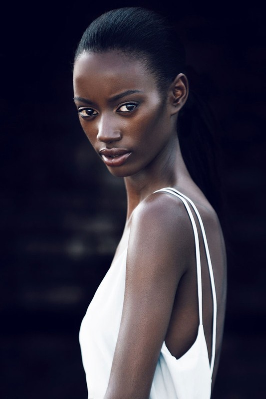 Portrait of a Black woman with dark hair pulled back in a ponytail, wearing a white dress against a dark blurred background.