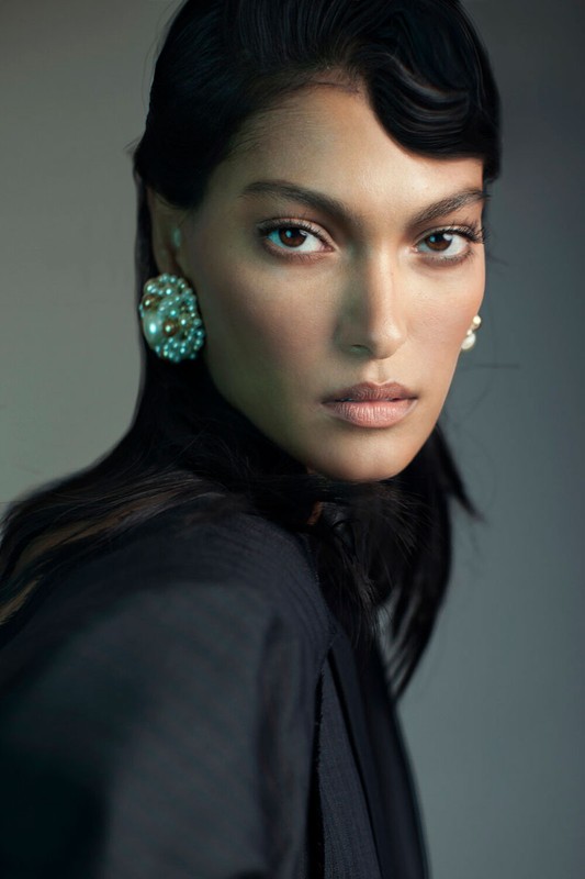 Close-up studio portrait of a woman with dark hair and pearl earrings wearing a black jacket with soft, diffused lighting.