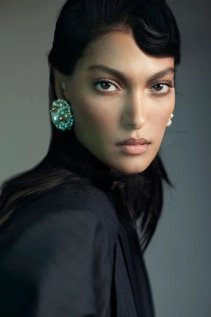 Close-up portrait of a woman with dark hair wearing pearl cluster earrings and a black blazer against a neutral grey background