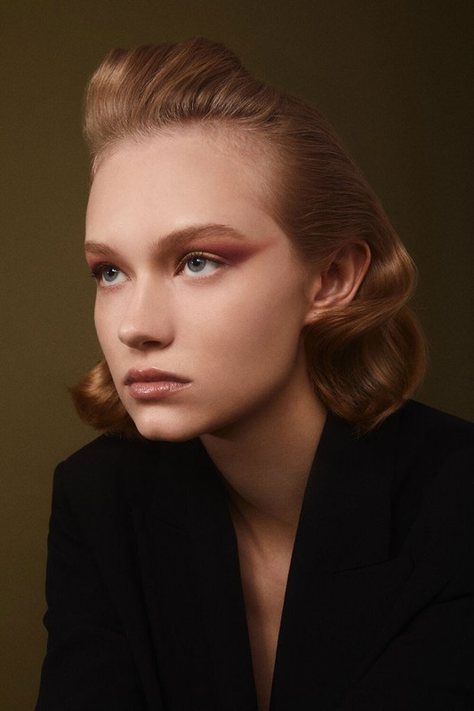 Portrait of a young woman with light brown hair and pink eye makeup, wearing a black blazer, against an olive green background.