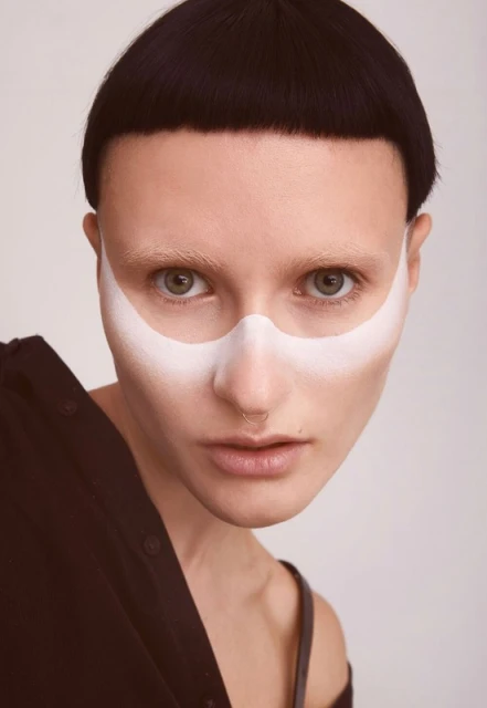 Close-up of a person with dark, blunt-cut hair and white face paint, wearing a dark top against a neutral background in a studio setting.