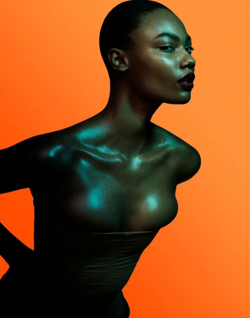 Portrait of a Black woman with dark red lipstick posing in front of an orange background with teal reflected light on her skin.