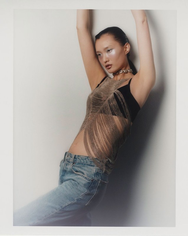Woman with arms raised wearing jeans and a fringed top in a studio setting. Fashion shot with soft lighting and a white background.