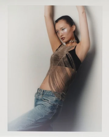 Woman with arms raised wearing jeans and a fringed top in a studio setting. Fashion shot with soft lighting and a white background.