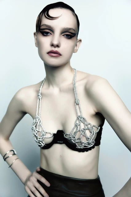 Close-up studio shot of a young adult woman with creative makeup and hairstyle wearing a silver mesh bra and bracelets against a white backdrop.