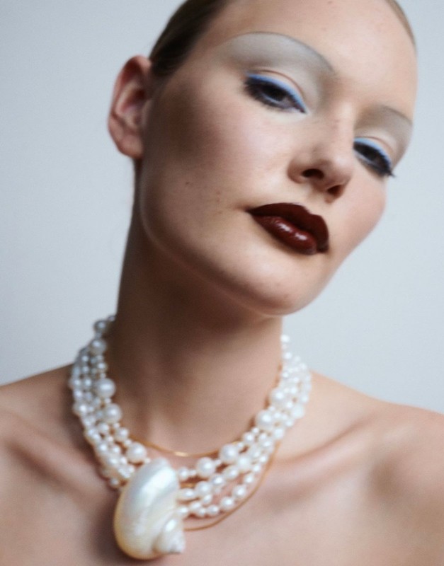 Adult female fashion model poses in studio wearing makeup and pearl necklace with shell pendant against a light background.
