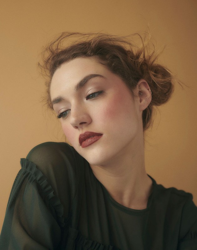 Close-up portrait of a woman with red lipstick and olive-green top against a warm, peach-colored background in soft, diffused lighting.