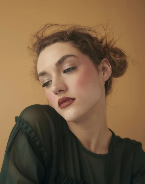 Close-up portrait of a woman with red lipstick and olive-green top against a warm, peach-colored background in soft, diffused lighting.