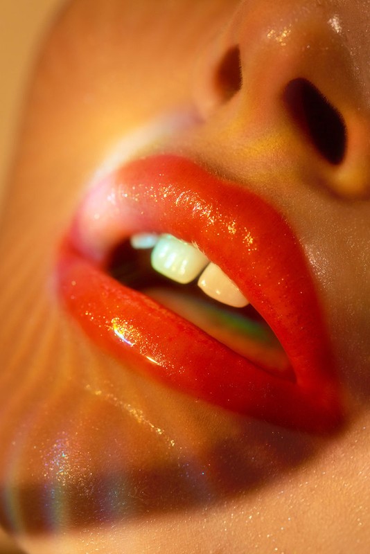Close-up macro shot of glossy red lips slightly parted with visible white teeth and warm rainbow light refraction on skin.