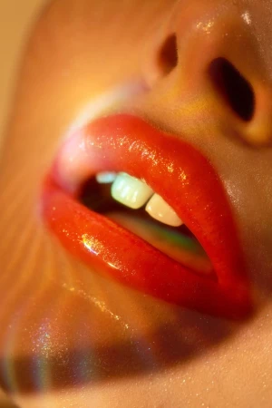 Close-up macro shot of glossy red lips slightly parted with visible white teeth and warm rainbow light refraction on skin.