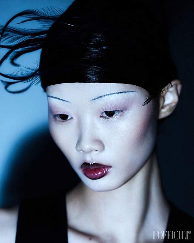 Close-up portrait of an Asian adult female fashion model with pale skin, red lips, and dramatic eye makeup, wearing black feathered headwear.