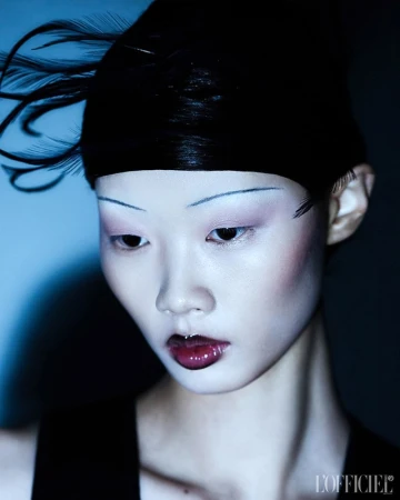 Close-up portrait of an Asian adult female fashion model with pale skin, red lips, and dramatic eye makeup, wearing black feathered headwear.