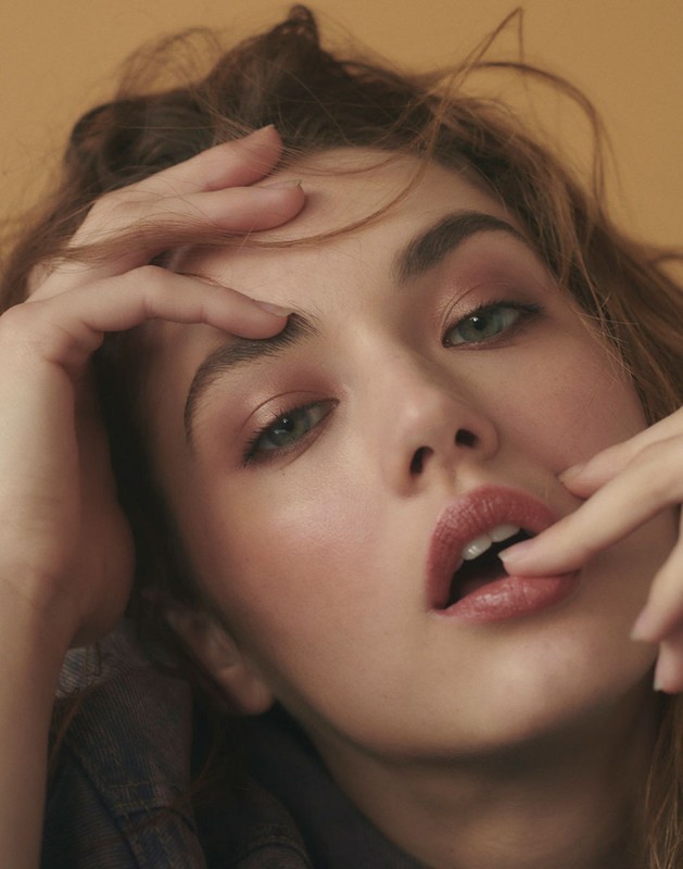 Close-up studio portrait of a young woman with fair skin, wearing a denim top, and light makeup while posing with her hands.