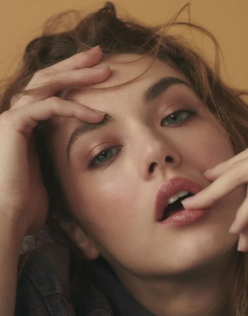 Close-up studio portrait of a young woman with fair skin, wearing a denim top, and light makeup while posing with her hands.