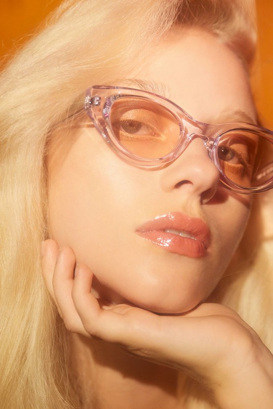 Close-up studio shot of a blonde woman wearing transparent lavender sunglasses, resting her face on her hand against an orange backdrop.
