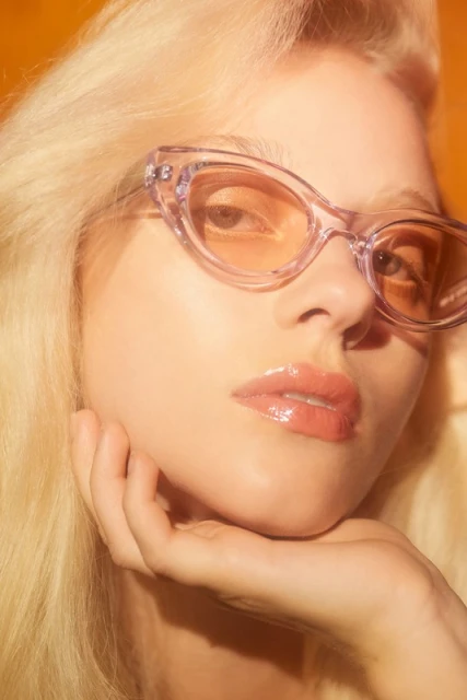 Close-up studio shot of a blonde woman wearing transparent lavender sunglasses, resting her face on her hand against an orange backdrop.
