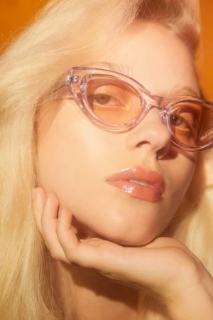 Close-up studio shot of a blonde woman wearing transparent lavender sunglasses, resting her face on her hand against an orange backdrop.