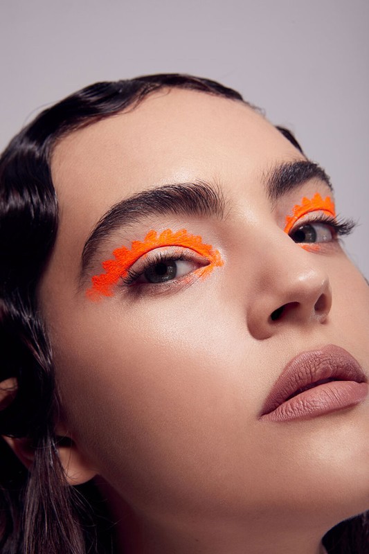 Close-up portrait of a young adult female with bright orange geometric makeup around her eyes, set against a neutral gray background.