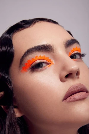 Close-up portrait of a young adult female with bright orange geometric makeup around her eyes, set against a neutral gray background.