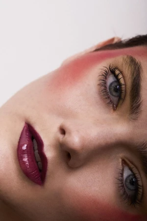 Close-up portrait of a fair-skinned woman with striking eye makeup, plum glossy lips, and a flushed pink blush in a studio setting.
