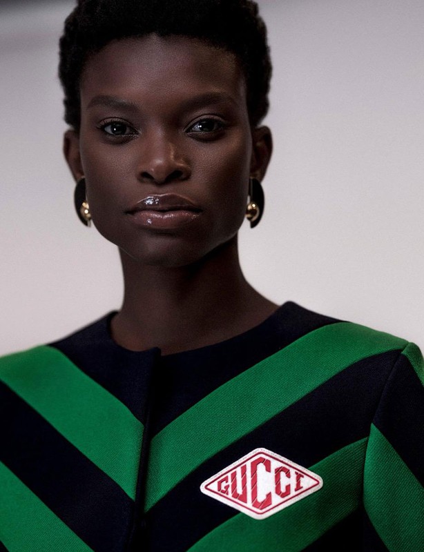 Close-up portrait of an adult female fashion model with dark skin and short dark hair wearing a green striped jacket with Gucci logo.