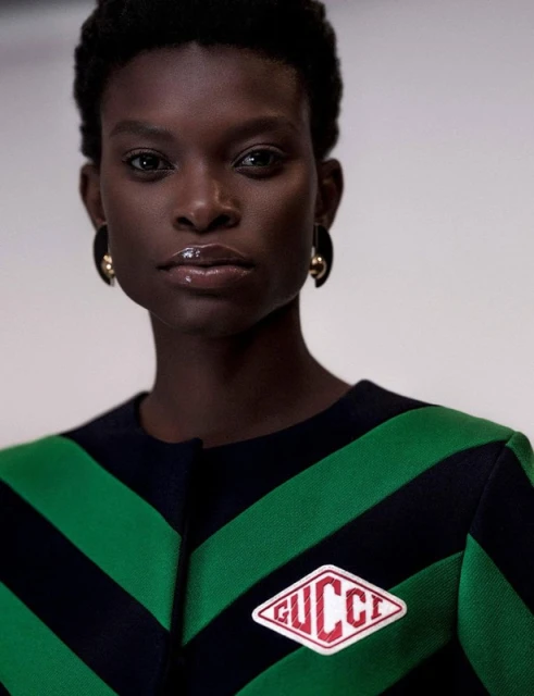 Close-up portrait of an adult female fashion model with dark skin and short dark hair wearing a green striped jacket with Gucci logo.