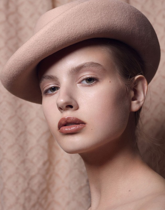 Close-up studio shot of a young woman wearing a beige hat and glossy lipstick, set against a textured neutral background.