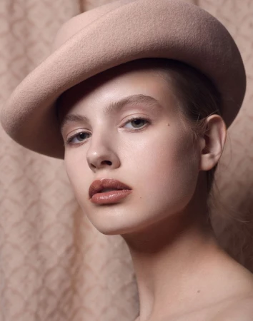 Close-up studio shot of a young woman wearing a beige hat and glossy lipstick, set against a textured neutral background.