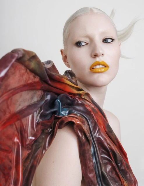 Headshot of a fair-skinned adult female fashion model with platinum blonde hair and yellow lipstick, wearing an artistic fabric shawl.