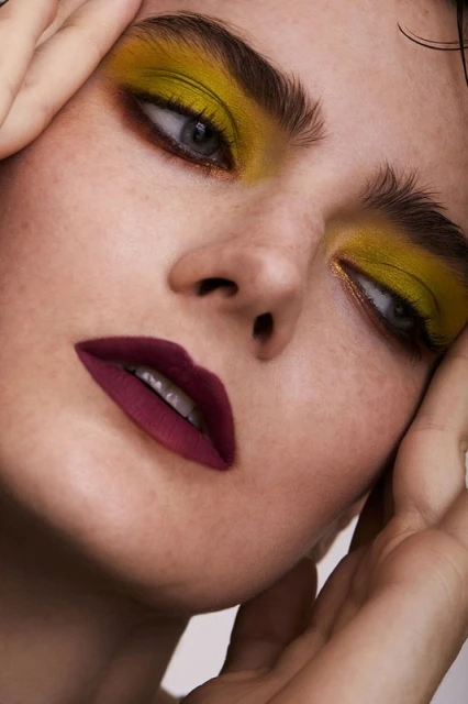 Close-up portrait of a woman with bold yellow eyeshadow and dark burgundy lipstick, hands framing her face in a beauty shot.