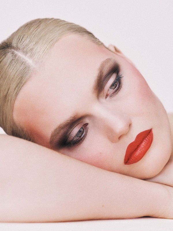 Close-up studio shot of a blond woman with bold red lipstick and dramatic eyeshadow, with her head resting on her arms against a white backdrop.