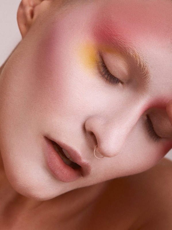 Close-up of a woman with closed eyes, colorful makeup on her face, and a nose ring, set against a light-colored background in a beauty portrait.