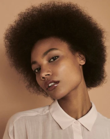 Portrait of a woman with a large afro hairstyle, wearing a light pink button-down shirt, against a light brown backdrop in a photo studio.