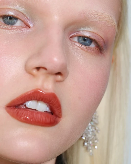 Close-up of a young adult female model with fair skin, light blonde hair, glossy lips, colorful eye shadow, and statement crystal earrings.