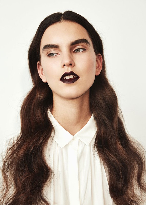 Portrait of a young woman with dark lipstick and wavy brown hair in a white shirt against a white background looking left.