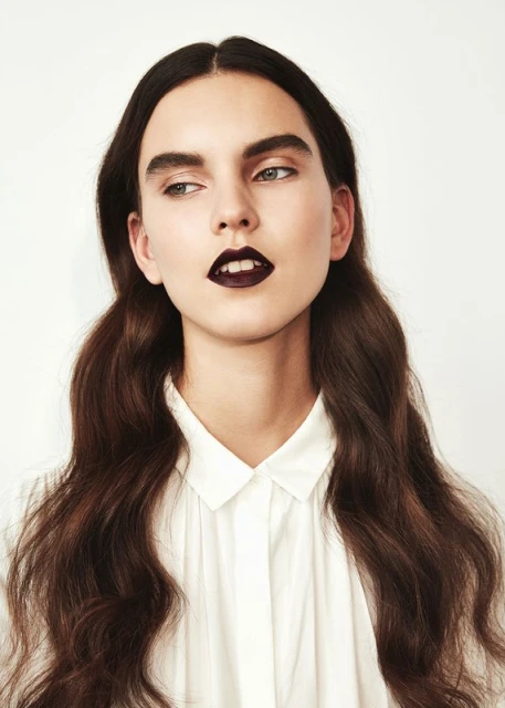 Portrait of a young woman with dark lipstick and wavy brown hair in a white shirt against a white background looking left.
