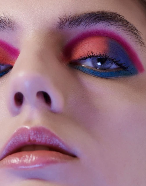 Close-up portrait of an adult female face with colorful eyeshadow in vibrant shades of pink, orange, blue, and teal.
