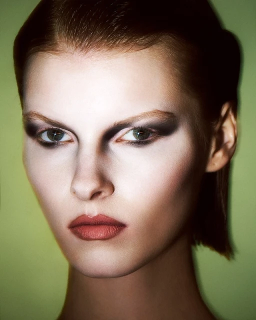 Close-up studio shot of a young fair-skinned adult female with dark eye makeup and pale lipstick against a muted green backdrop.