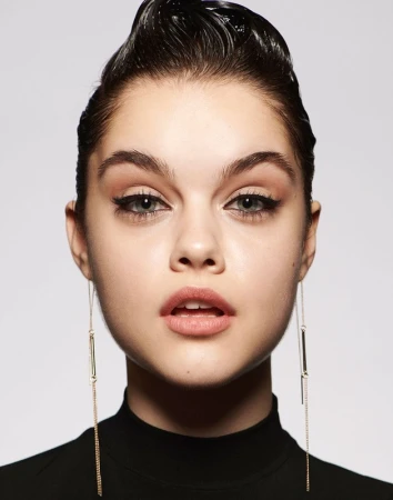 Close-up portrait of a woman with slicked-back dark hair, winged eyeliner, and long drop earrings against a neutral grey background.
