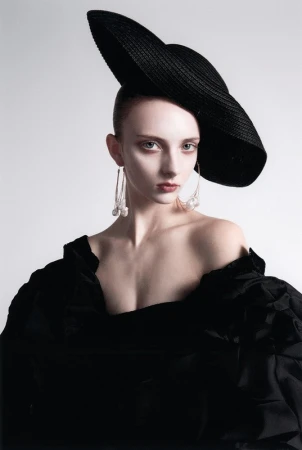 Model wearing a large black tilted hat, off-the-shoulder black ruffled top, and long drop earrings against a plain white background