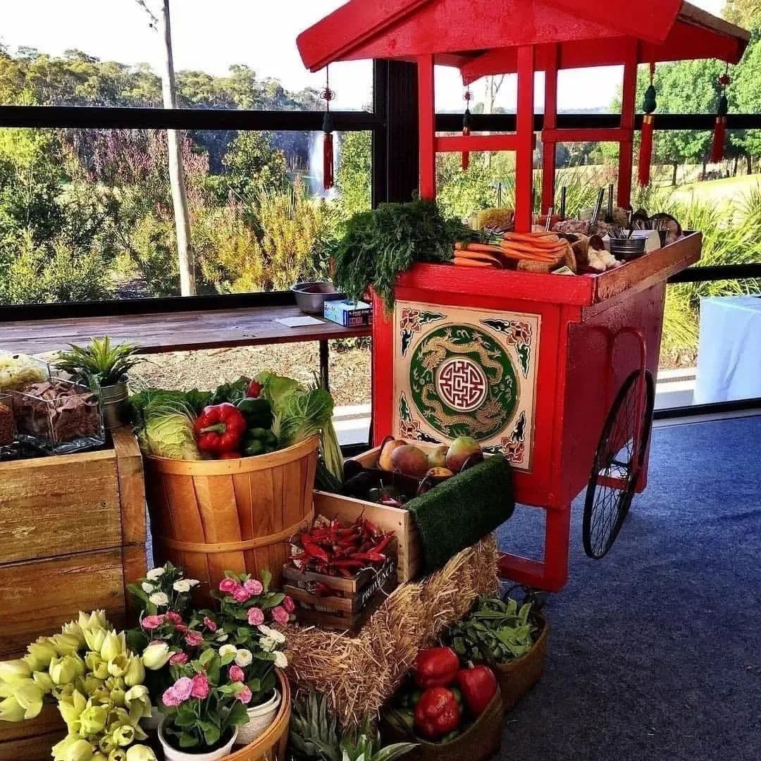 A red vendor cart with Asian design is surrounded by fresh produce like lettuce and peppers, outdoors with soft natural light.