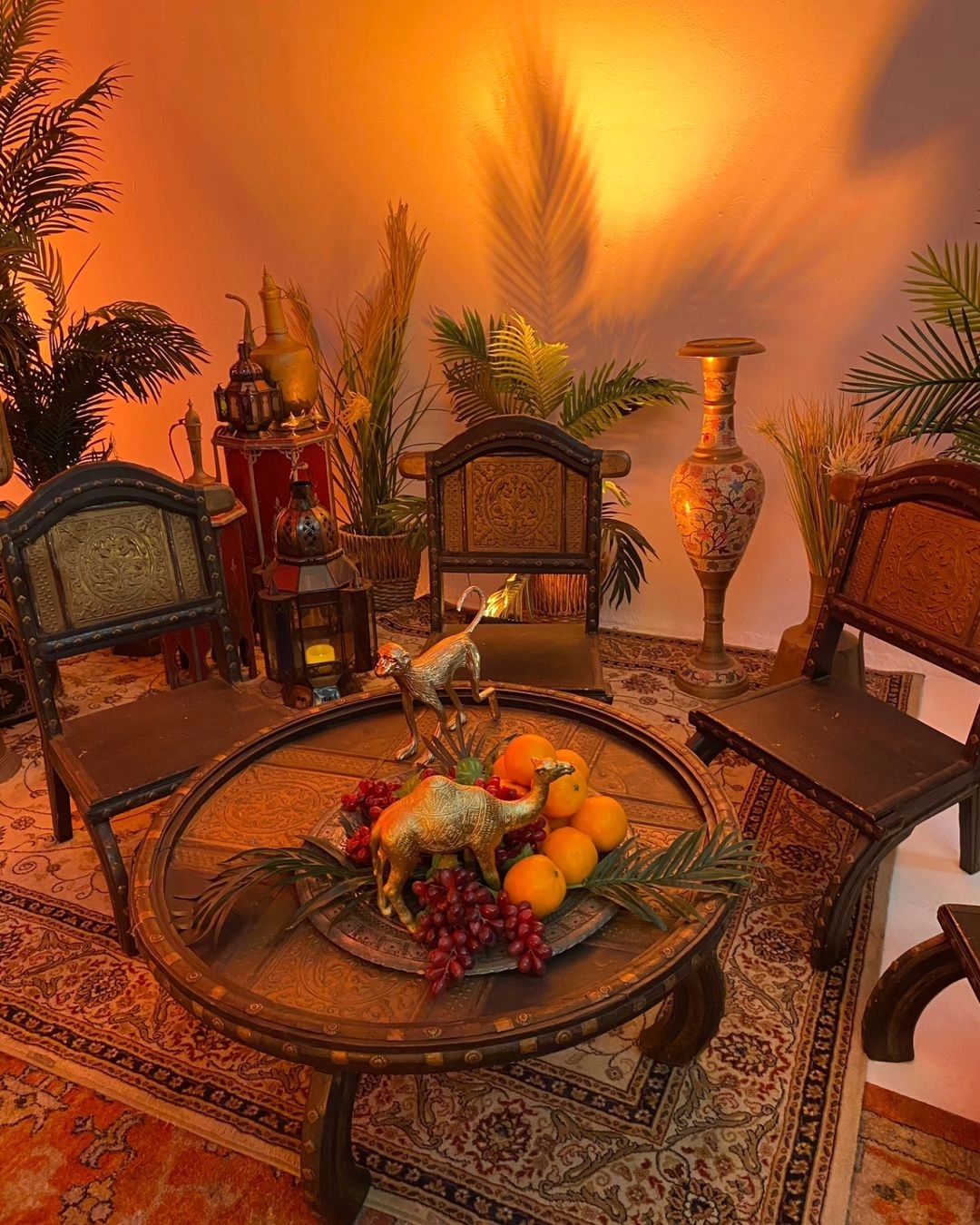 Ornate round table with golden camel figurines, grapes, and oranges, surrounded by chairs, potted plants, and carpet, in a warm room.