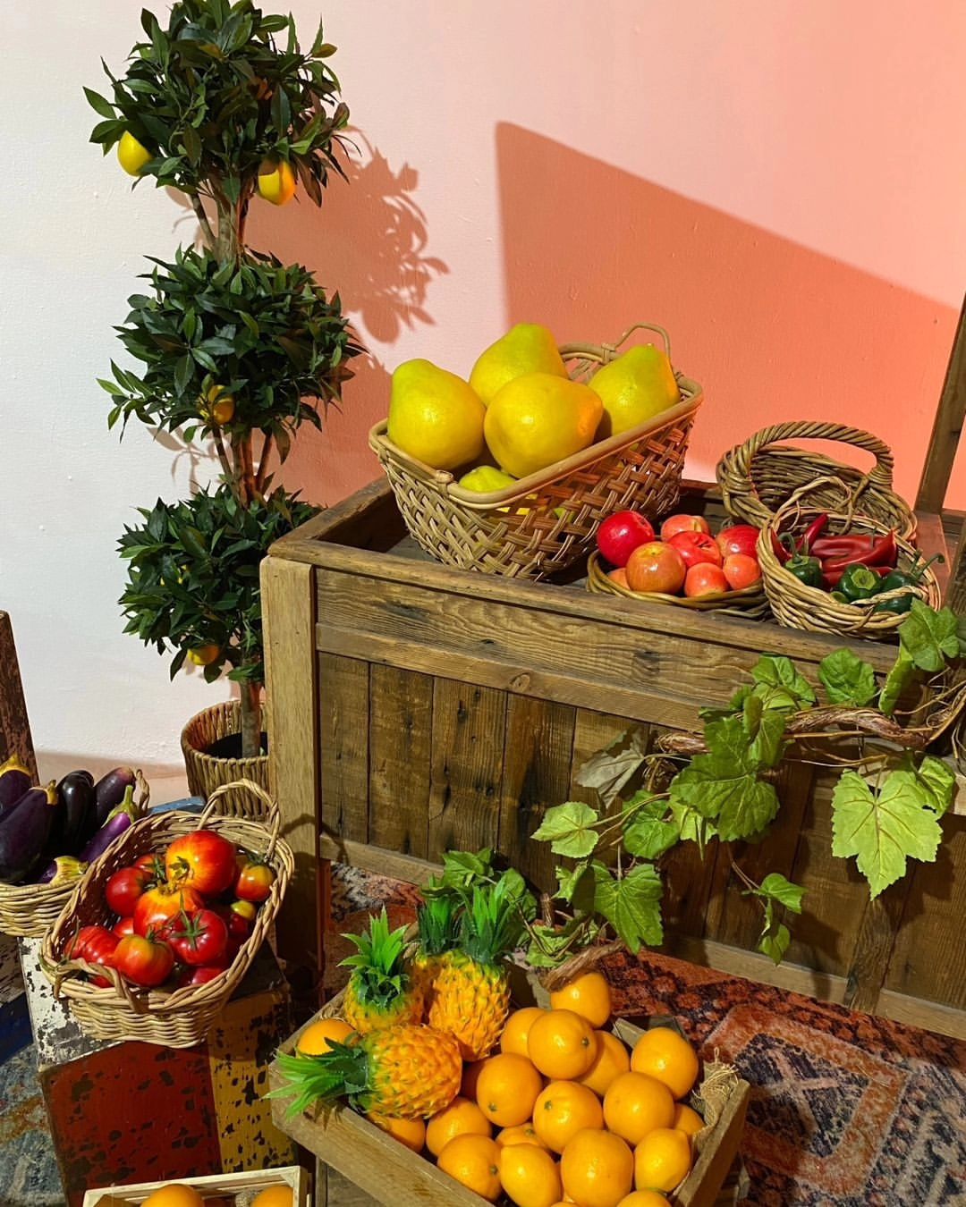 Baskets of vibrant fruits and vegetables artfully arranged on a rustic wooden table next to a small fruit tree against a muted pink wall.