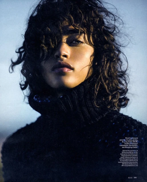 Headshot of a female fashion model with curly dark hair, wearing a black knit turtleneck sweater in front of an outdoor background.