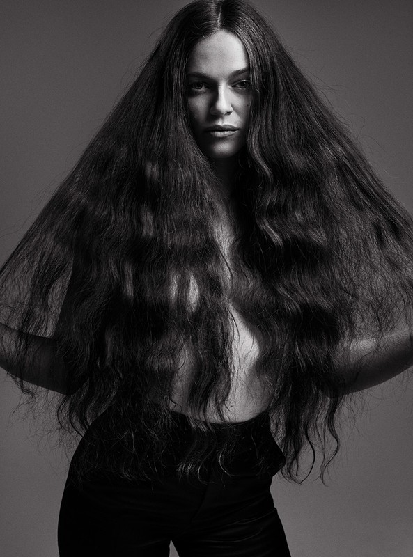 Portrait of a woman with long, dark wavy hair, posing in black pants against a gray background in a studio.