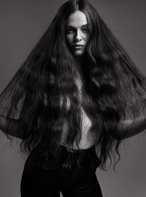 Portrait of a woman with long, dark wavy hair, posing in black pants against a gray background in a studio.