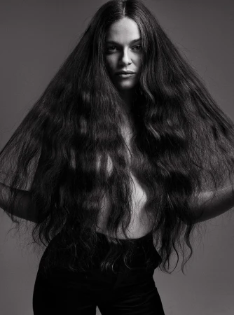 Portrait of a woman with long, dark wavy hair, posing in black pants against a gray background in a studio.